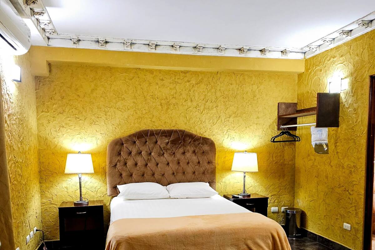 A room with a large bed, two bedside tables with lamps, a wall-mounted shelf, and a trash bin.