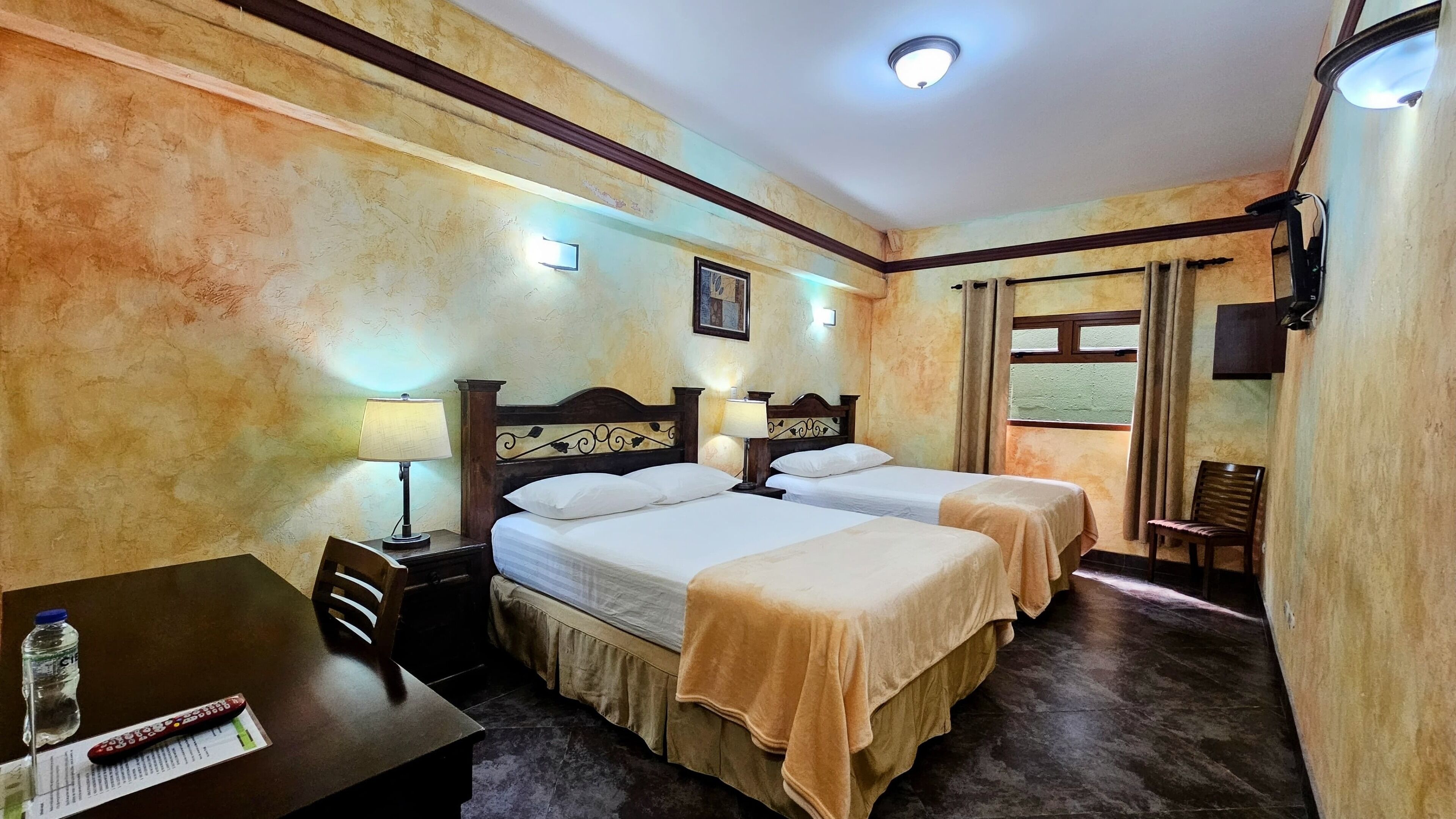 A hotel room with two beds, a desk with a chair, a table with a bottle and remote control, and a window with curtains.