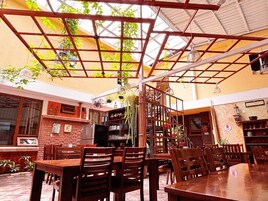 An indoor dining area with wooden tables and chairs, a brick wall with framed pictures, and a ceiling with a grid pattern.