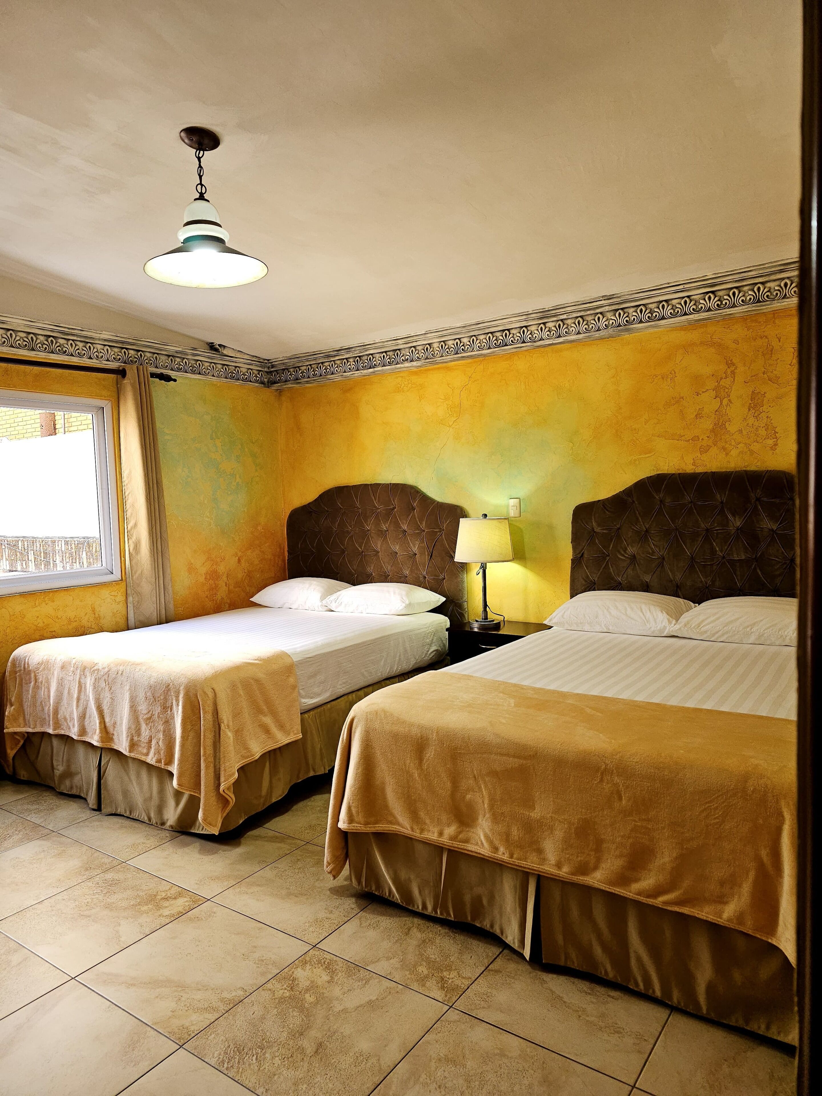 Une chambre d’hôtel avec deux lits simples, une table de chevet avec une lampe, une fenêtre avec des rideaux et un luminaire au plafond.
