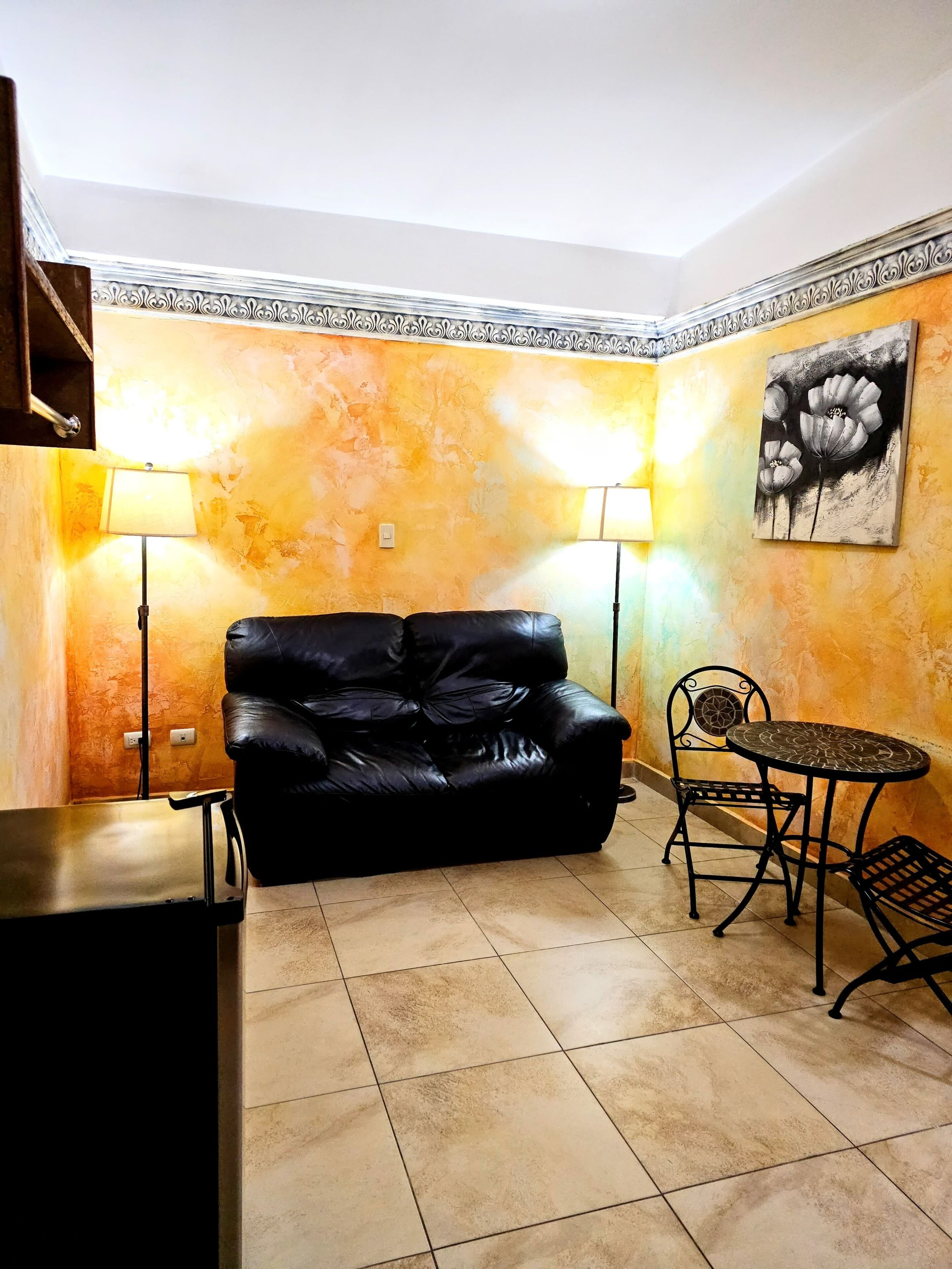 A living room with a black leather sofa, a round metal table, two chairs, and a wall-mounted lamp.