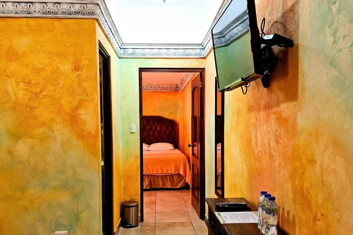A hotel room with a bed, a TV mounted on the wall, a small table with a water bottle, and a trash bin.