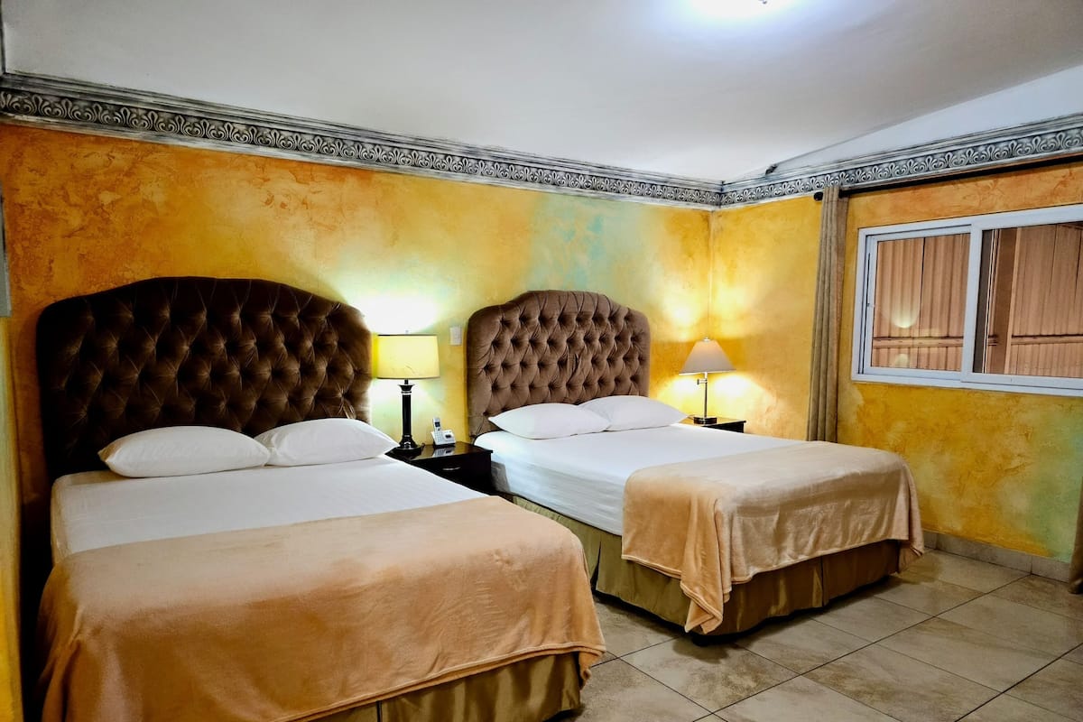 A hotel room with two beds, each with a tufted headboard, a bedside table with a lamp, and a window with curtains.