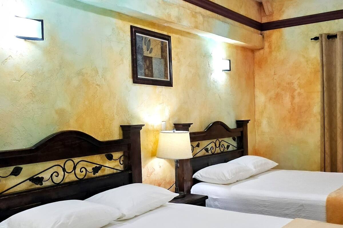A hotel room with two beds, a nightstand with a lamp, a painting on the wall, and a window with curtains.