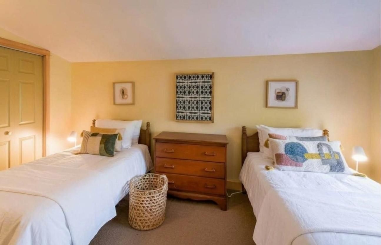 Une chambre avec deux lits simples, une commode en bois, un panier en osier et des tableaux encadrés accrochés au mur.