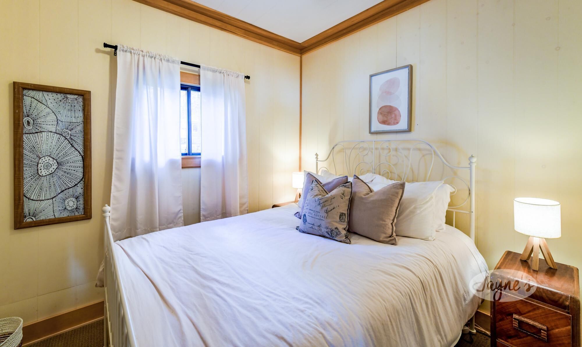 Un dormitorio con cama, mesita de noche, lámpara, ventana con cortinas y dos cuadros en la pared.