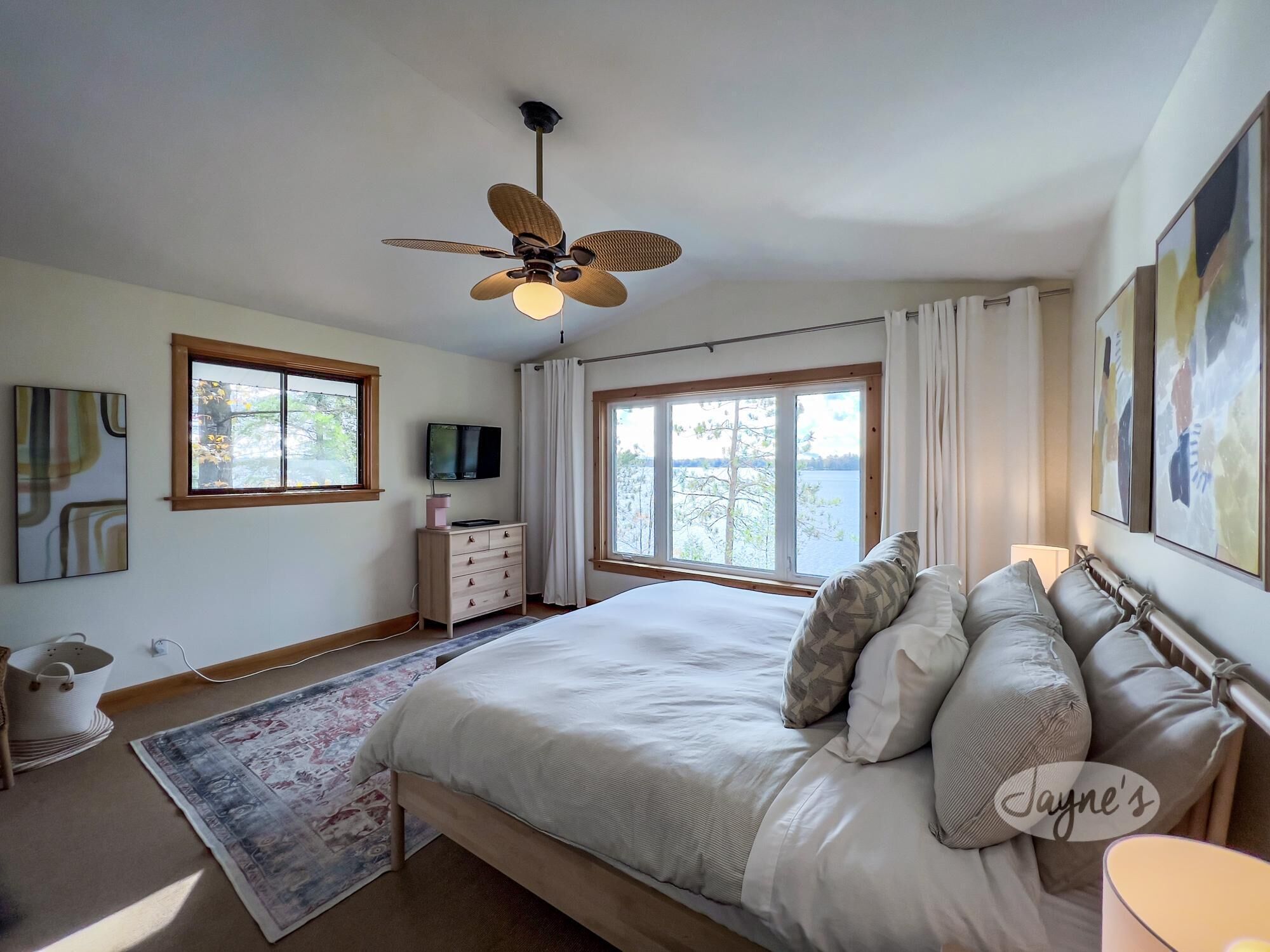 Un dormitorio con cama, ventilador de techo, cómoda, televisor y dos ventanas con vistas a un lago.