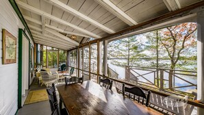 Un espacio comedor techado con una mesa y sillas de madera, ventanas amplias con vistas a un lago y árboles, y un techo de madera con vigas a la vista.