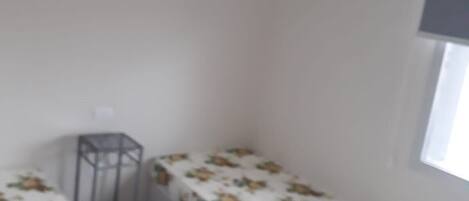 A small, sparsely furnished room with two single beds, a small table, and a window.