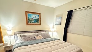 A neatly made bed with a quilted cover, a ceiling fan, a nightstand with a lamp, and a painting on the wall.