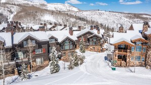 Un chalet enneigé composé de plusieurs bâtiments, avec une piste de ski en arrière-plan et un ciel dégagé.