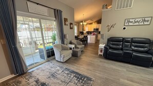 A spacious living room with a black leather sofa, a striped armchair, and a grey armchair. There is a large rug on the floor, and a sliding glass door leading to a patio. The kitchen is visible in the background with white cabinets and a hanging light fixture. - Pelican Landing / Beach Bird Properties - Inside Oceanside Village (Surfside Beach)