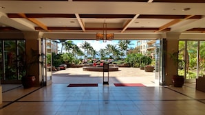 Un hall d’entrée spacieux avec un sol carrelé, un espace de détente central agrémenté de coussins rouges, et une vue sur des palmiers et une piscine.