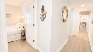 A hallway with a bed, a nightstand, a lamp, a wall-mounted mirror, and a door leading to another room.