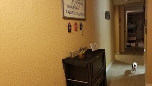 Un pasillo con una pared beige texturizada, una cita inspiradora enmarcada, una pequeña casa decorativa y un perro a lo lejos.