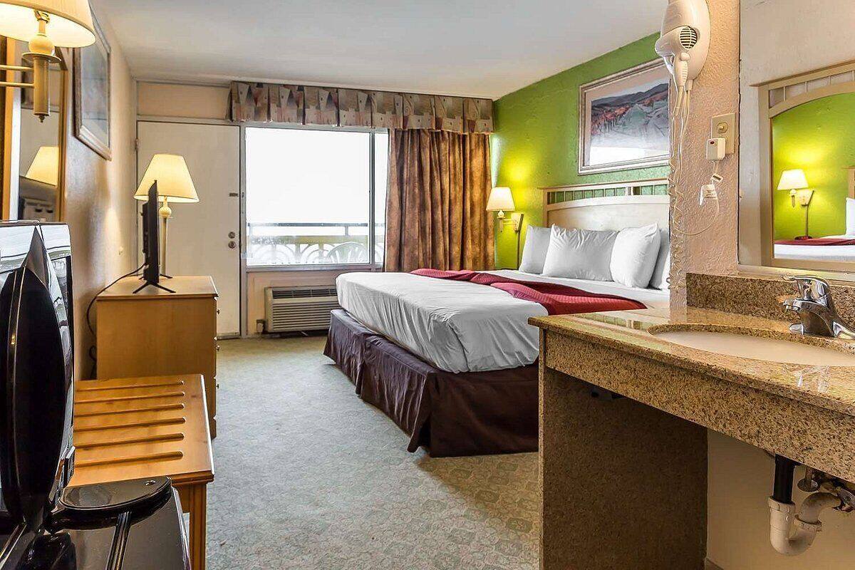 A hotel room with a bed, a desk with a lamp, a mirror, and a window with curtains.