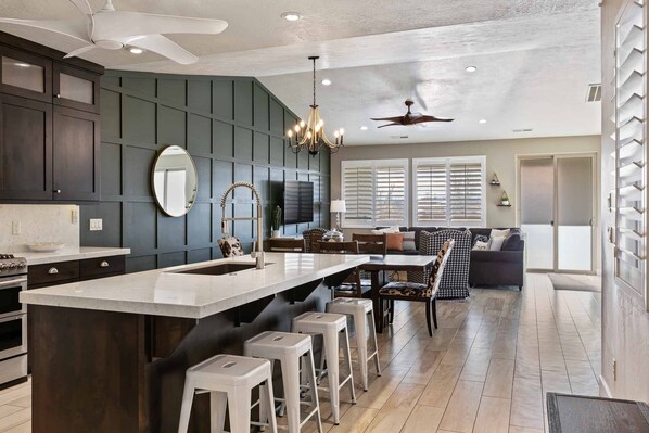 A modern kitchen with dark cabinetry, a large island with stools, a ceiling fan, and a dining area with a sofa and chairs.