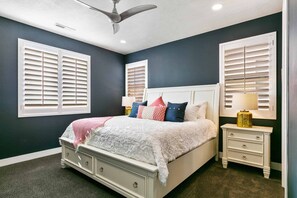 A bedroom with a bed, two nightstands, a ceiling fan, and two windows with blinds.