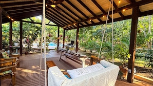 A covered outdoor seating area with a swing chair, wooden benches, and a pool in the background.
