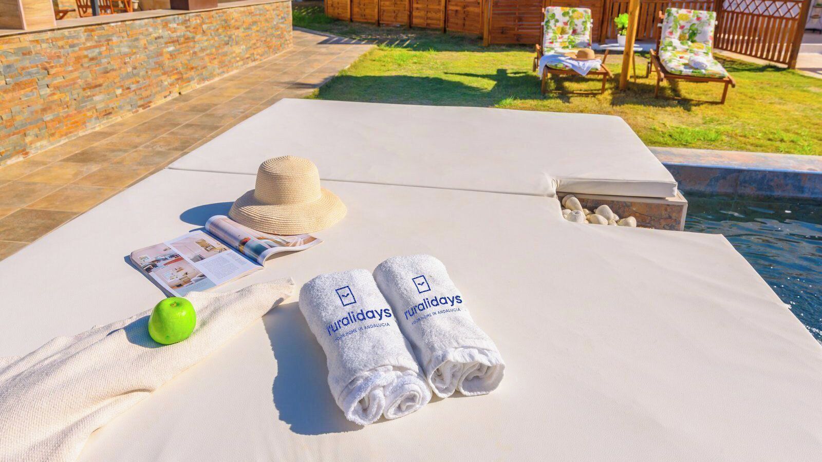 Zona de piscina con una silla de descanso blanca, un sombrero de paja, una revista, dos toallas enrolladas y una manzana verde sobre una toalla.