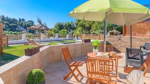 An outdoor patio with a table, chairs, and a large umbrella. There are potted plants and a wooden fence in the background.