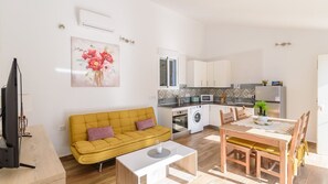 A modern living room with a yellow sofa, a dining table with chairs, a TV, and a kitchen area with cabinets, a microwave, and a washing machine.