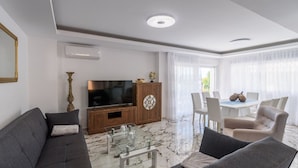 Een moderne woonkamer met een flatscreen televisie, een houten tv-meubel, een glazen salontafel en een eethoek met witte stoelen en een tafel.