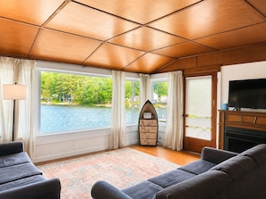 A living room with a large window, a fireplace, a TV, and a surfboard-shaped Wi-Fi sign. - Quiet Lakefront Cabin · Private Dock and Firepit (Bath)