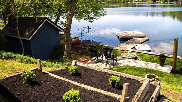Una cabaña junto al lago con un muelle, un cobertizo pequeño y un jardín con bancales elevados.
