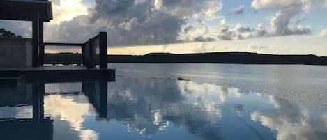 A serene infinity pool reflecting the sky and clouds, with a modern structure and a view of the distant shoreline.