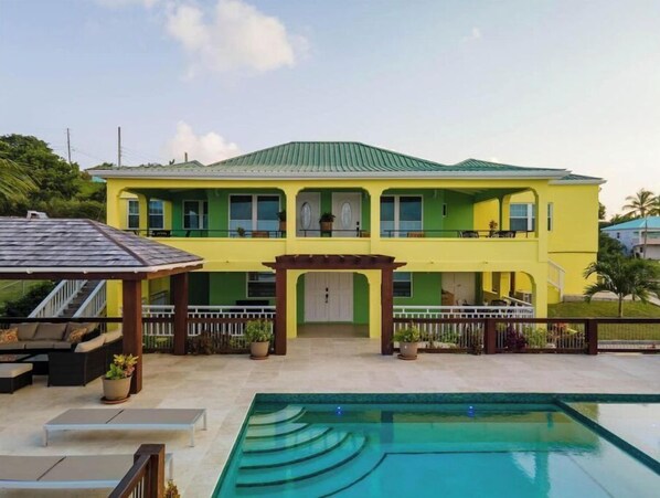 Ein gelbes Haus mit zwei Stockwerken, einem grünen Dach, einem Pool und einer überdachten Terrasse.