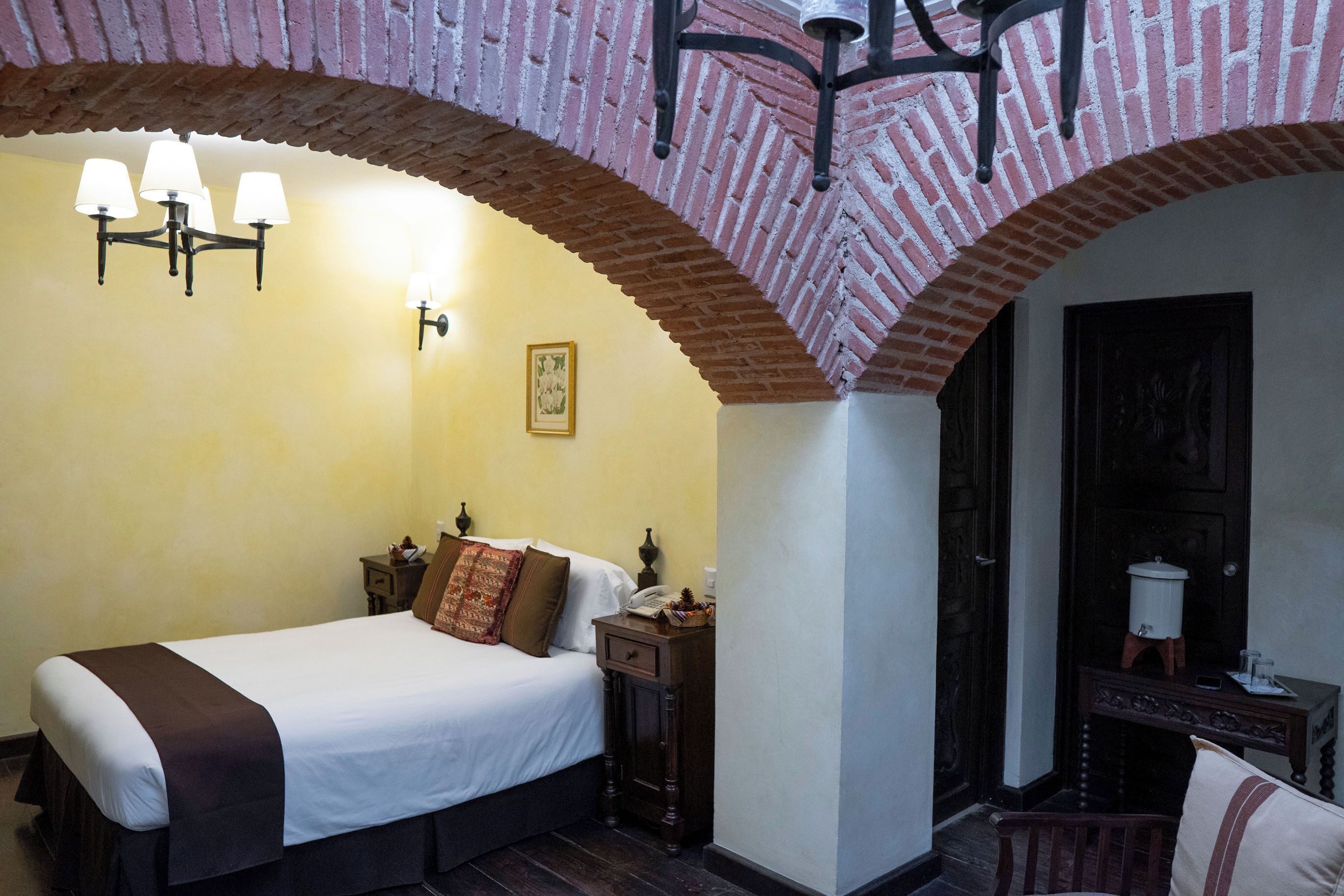 Quarto de hotel com cama, mesa de cabeceira, luminária, quadro na parede e uma porta.