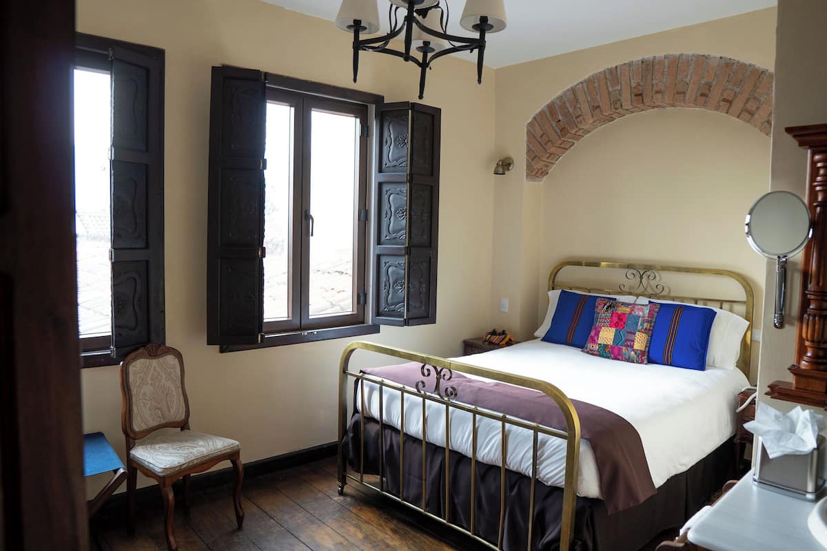 Ein Schlafzimmer mit einem Metallbettgestell, einem Holzstuhl, einem kleinen Tisch und einem Fenster mit Fensterläden.