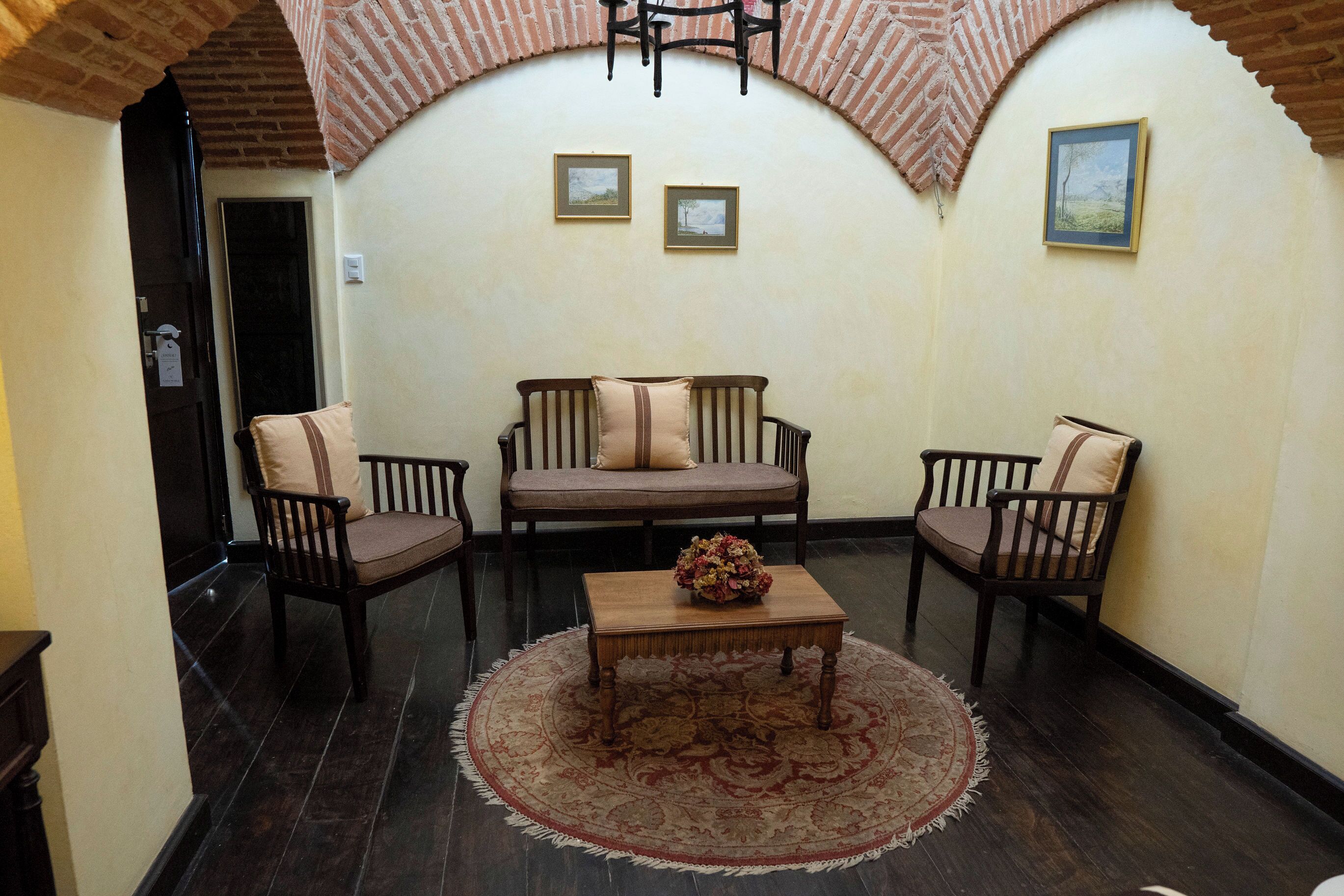 A cozy room with a dark wooden floor, a round rug, a wooden coffee table with a floral centerpiece, and two wooden chairs with cushions.