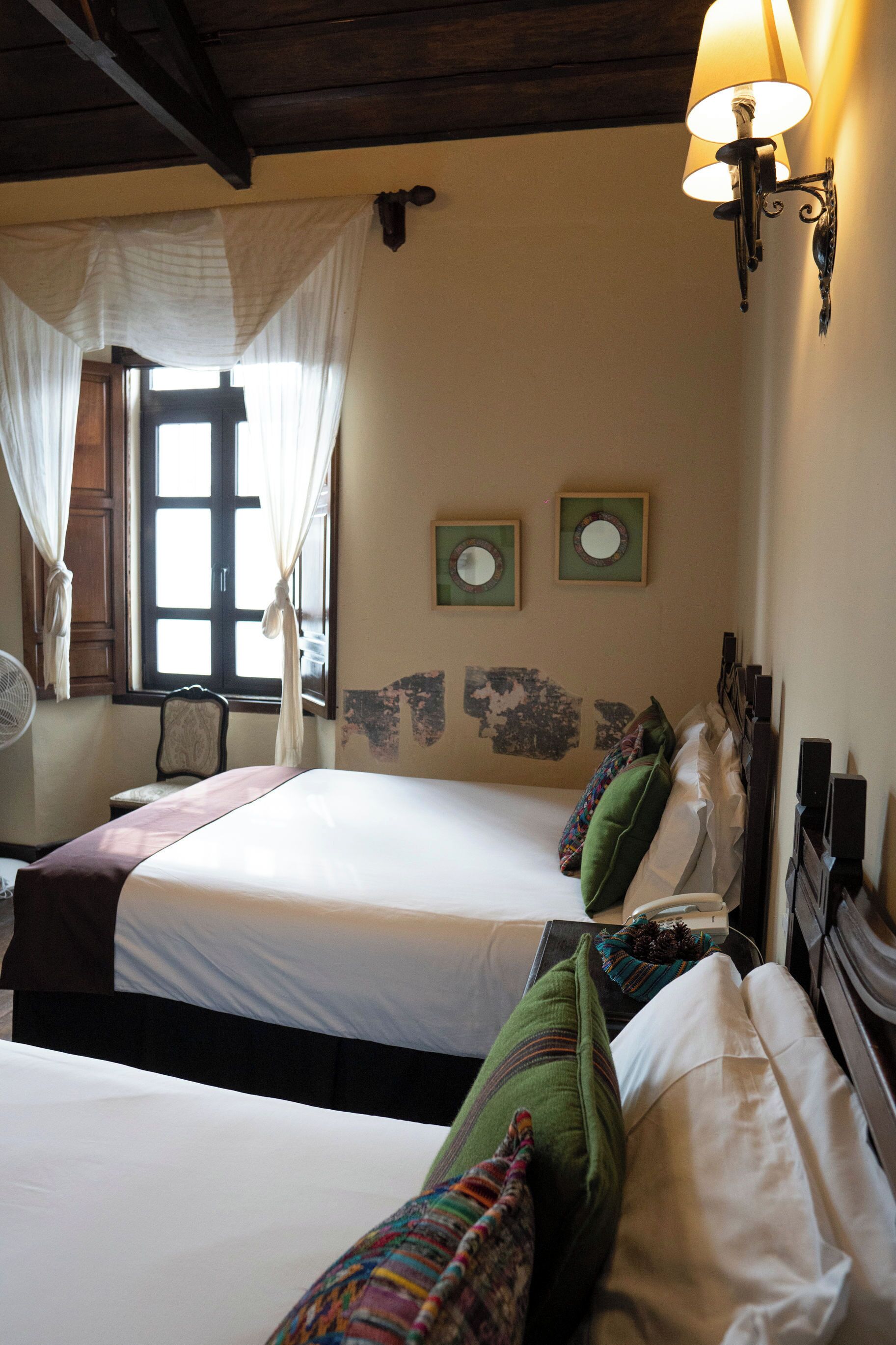 A room with two beds, a window with curtains, a wall-mounted lamp, and a chair.