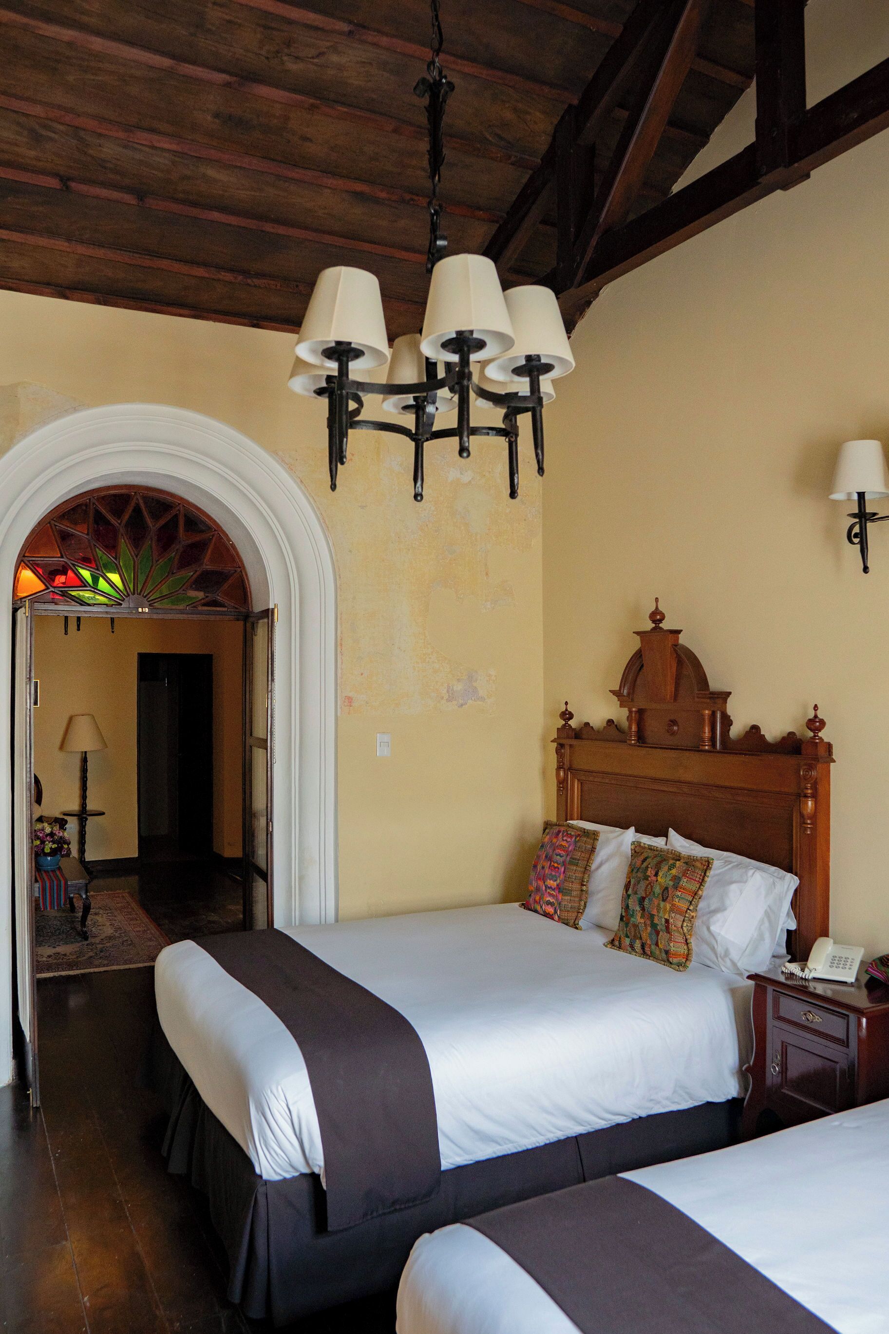 Habitación de hotel con dos camas, cabecero de madera, candelabro, teléfono y puerta que da a otra habitación.