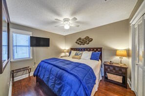 A bedroom with a bed, a ceiling fan, a TV mounted on the wall, and a bedside table with a lamp.