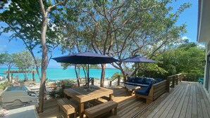 A wooden deck with outdoor furniture, including a table and benches, overlooking a beach with clear blue water.