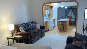 A cozy living room with a sofa, ottoman, and a small table with a lamp. A kitchen and dining area are visible through an open archway.