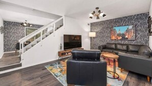 Una sala de estar moderna con un sofá de cuero negro, un televisor de pantalla plana, una escalera y un cuadro de un paisaje urbano en la pared.
