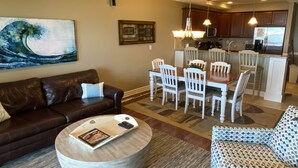A living room with a brown leather sofa, a round coffee table with magazines, a patterned armchair, and a dining area with white chairs and a wooden table.