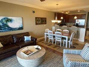 A living room with a brown leather sofa, a round coffee table with magazines, a patterned armchair, and a dining area with white chairs and a wooden table.