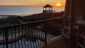 A balcony with a view of the sunset over the ocean, a pool, and outdoor seating.