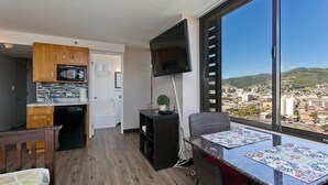 A modern kitchen with wooden cabinets, a built-in microwave, and a dishwasher. There is a dining area with a table and chairs, and a large window offering a city view. - 🗝18th Floor Corner Studio of Hawaiian Monarch w/Awesome Pool in Waikiki (Honolulu)