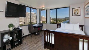 A hotel room with a large TV, a desk with a chair, a bed, and a view of a cityscape through large windows. - 🗝18th Floor Corner Studio of Hawaiian Monarch w/Awesome Pool in Waikiki (Honolulu)