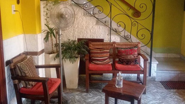 A room with a yellow wall, a staircase with a decorative railing, a fan, and a seating area with chairs and a table. - Bambi (La Falda)