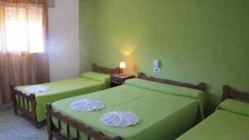 Una habitación con tres camas individuales, cada una con una colcha verde y una toalla blanca sobre la cama. Hay una mesita de noche con una lámpara entre las camas.