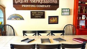 A dining area with a wooden table, black chairs with red cushions, a ceiling fan, and wall decorations including a 'BAR' sign, a 'Great Impressions Printing Company' sign, and a 'Picnic Ground No Overnight Camping' sign. -  Spacious- 11 min to Yosemite toll- 13 guests- 7+ beds, pet- game room- 2000 Ft (Oakhurst)