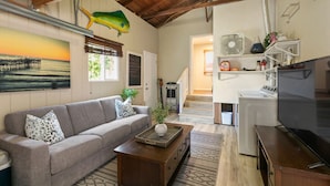 A cozy living room with a sofa, a coffee table, and a TV. There's a staircase leading to another room, and a large fish decoration on the wall. - Felspar House · Clean, Charming House in PB 🏖☀️ AC, pkg & laundry (San Diego)