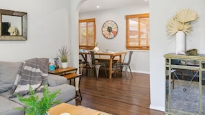 A living room with a gray sofa, a coffee table, and a dining area with a wooden table and chairs. There is a clock on the wall and a decorative vase on the sideboard. - Felspar House · Clean, Charming House in PB 🏖☀️ AC, pkg & laundry (San Diego)